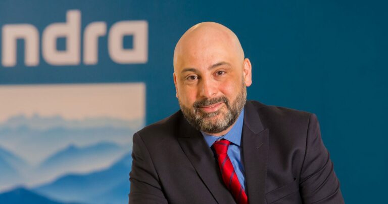 Marcelo Bernardino, CEO da Minsait no Brasil, está posicionado em frente a um fundo azul com o logotipo da empresa Indra. Ele veste um terno preto, camisa azul e gravata vermelha listrada, e exibe um sorriso sutil. A imagem transmite uma atmosfera profissional e corporativa