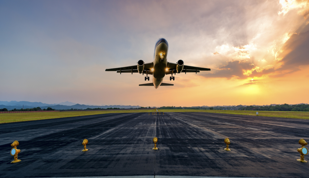 Avião decolando em uma pista de aeroporto ao pôr do sol, com o céu em tons alaranjados e montanhas ao fundo. A cena transmite movimento e a grandiosidade da aviação, aereo avião, avião decolando, decolagem, turismo