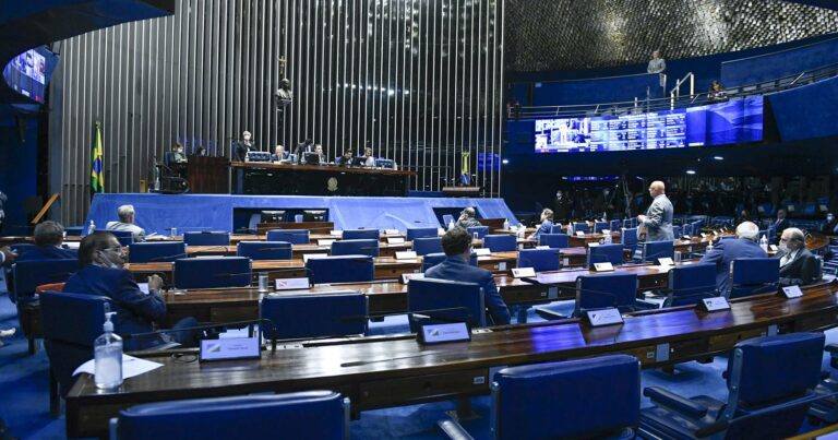 Plenário do Senado Federal do Brasil durante uma sessão, com senadores presentes em suas cadeiras dispostas em formato semicircular. No centro, a mesa diretora conduz os trabalhos sob a bandeira nacional e o brasão da República. O ambiente é iluminado, com painéis digitais ao fundo exibindo informações (Senado, plenário, projeto)