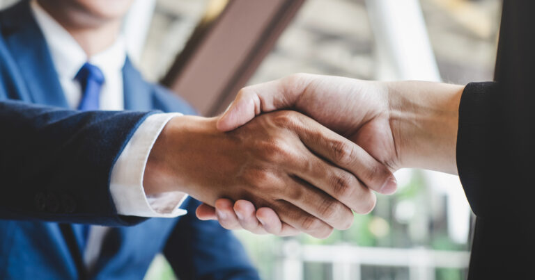 Close de um aperto de mãos entre duas pessoas vestidas formalmente, representando uma negociação de negócios, fusões e aquisições (M&A). Ao fundo, um ambiente corporativo com luz natural entra através das janelas (aquisição, compra, venda, negócios, TE Connectivity, topaz, CADE aprova, dimensa, totvs, Netwrix, cohesity, Udacity, Zucchetti, minsait)