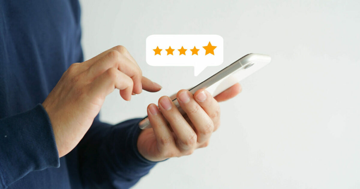 Imagem de uma pessoa segurando um smartphone com uma das mãos enquanto utiliza a outra para tocar na tela. Acima do dispositivo, há um balão de diálogo com cinco estrelas douradas, representando uma avaliação de alta qualidade ou feedback positivo. O fundo é branco, destacando a interação com o aparelho (clientes, experiência do cliente, CX)