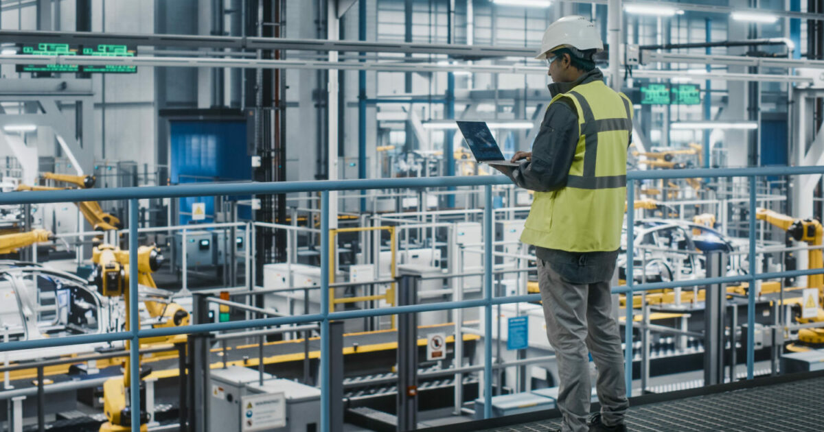 Engenheiro de manufatura em uma fábrica automatizada, utilizando um laptop para monitorar processos industriais. Ele veste colete refletivo e capacete de segurança, observando a linha de produção repleta de braços robóticos montando veículos. O ambiente é moderno e altamente tecnológico, representando a automação e a digitalização na indústria (dispositivos)