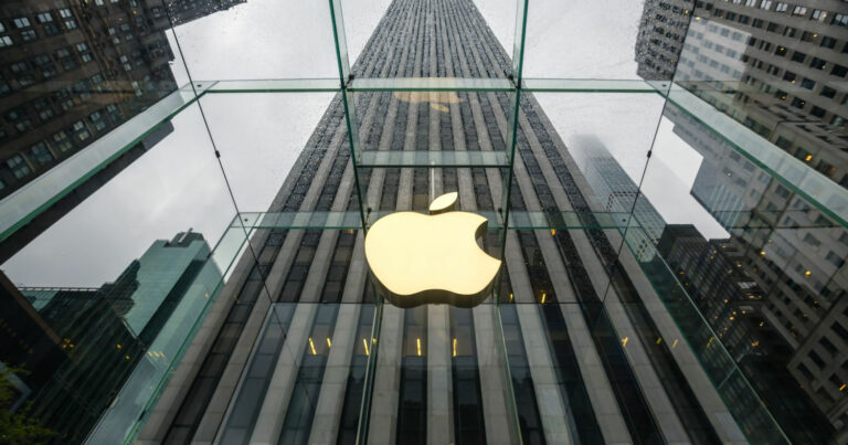 Fachada de uma loja da Apple com o icônico logotipo iluminado em destaque no centro de uma estrutura de vidro. O ângulo da imagem oferece uma perspectiva imponente, mostrando edifícios altos ao fundo e um céu nublado, ressaltando a modernidade e sofisticação da marca (apple, lançamento, inovação)
