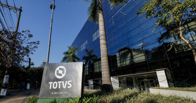 Fachada do prédio da TOTVS, com estrutura moderna de vidro refletindo o céu e as árvores ao redor. Em primeiro plano, destaca-se uma placa com o logotipo da TOTVS e o endereço 'Av. Braz Leme, 1000'. O cenário é complementado por palmeiras e vegetação, transmitindo um ambiente corporativo sofisticado e integrado à natureza (Totvs VTEX Linx)