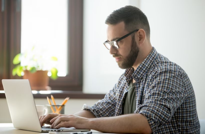 Homem, de óculos e barba, vestindo camisa xadrez em um escritório, de perfil, mexendo no computador.