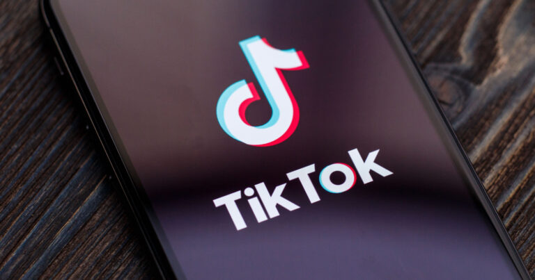 Ícone do aplicativo TikTok exibido na tela de um smartphone sobre uma superfície de madeira escura. O logotipo característico do TikTok aparece com suas cores neon em azul e rosa, contrastando com o fundo preto do dispositivo, representando o uso de redes sociais em dispositivos móveis (Latam, aviões, nebraska)