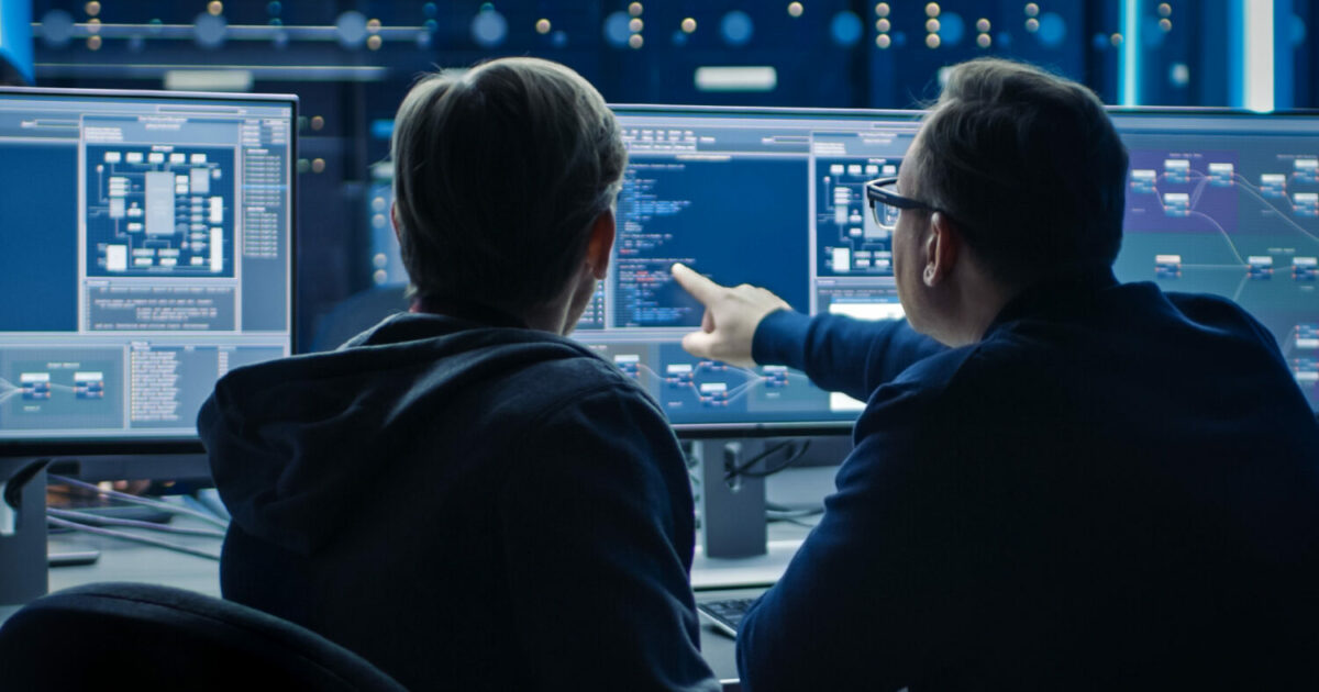 Dois profissionais de cibersegurança analisando códigos e gráficos em múltiplos monitores em um ambiente tecnológico. Um deles aponta para a tela, indicando uma possível solução ou análise crítica. Representação do trabalho de CISOs e especialistas em segurança digital (CISOs, cibersegurança, incidentes de segurança, empresas)