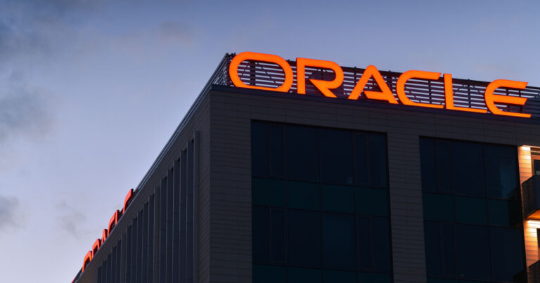 Fachada de um prédio da Oracle ao entardecer, com o logotipo da empresa iluminado em vermelho no topo do edifício. O céu ao fundo está parcialmente nublado, destacando a arquitetura moderna do local (nuvem, Donald Trump)
