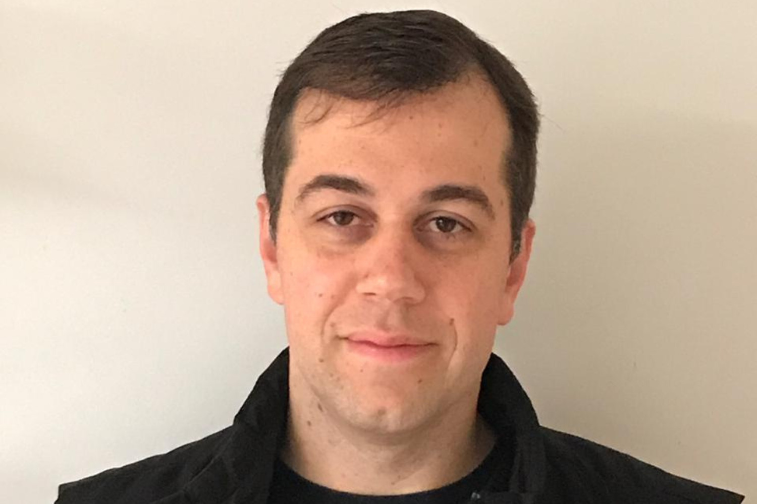 “Pessoa com cabelos curtos e castanhos, vestindo uma jaqueta preta sobre uma camiseta escura, posicionada contra um fundo branco (liderança)