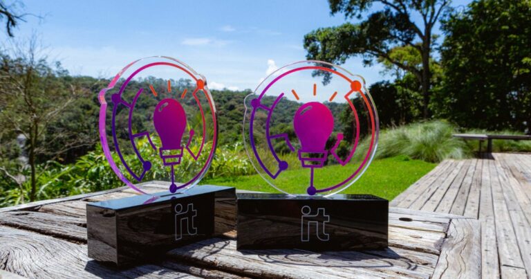 Troféus do prêmio 'As 100+ Inovadoras no Uso de TI' expostos ao ar livre sobre uma mesa de madeira. Os troféus têm formato moderno com um design de lâmpada estilizada em tons de roxo e laranja, destacando-se contra um cenário de natureza verde com árvores e céu azul ao fundo, IT Forum