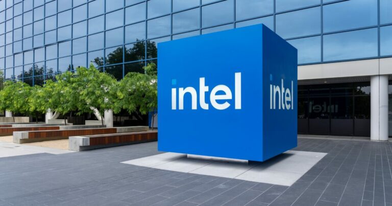 Logotipo da Intel em um grande cubo azul localizado na entrada de um edifício corporativo moderno, com fachada de vidro reflexivo e árvores ao fundo. O espaço ao redor conta com bancos e áreas de convivência