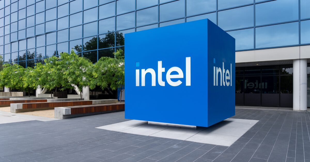 Logotipo da Intel em um grande cubo azul localizado na entrada de um edifício corporativo moderno, com fachada de vidro reflexivo e árvores ao fundo. O espaço ao redor conta com bancos e áreas de convivência