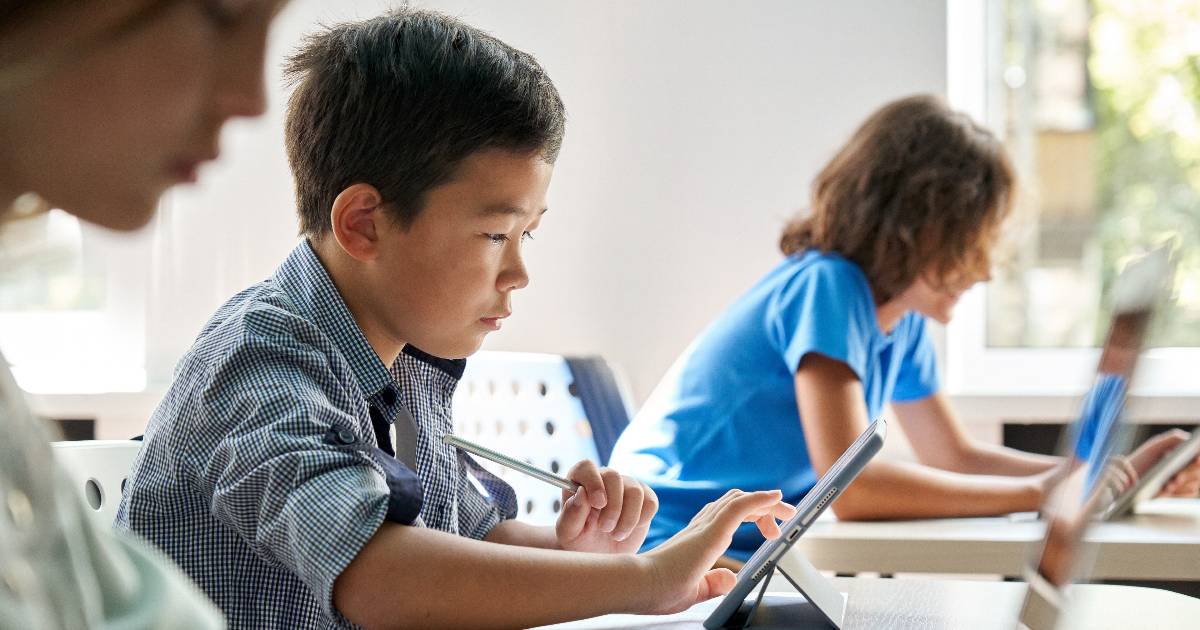 Grupo de crianças em uma sala de aula moderna, concentradas em atividades usando tablets. Em primeiro plano, um menino com expressão focada toca a tela do tablet com uma caneta digital, enquanto outras crianças ao fundo também interagem com dispositivos tecnológicos. A luz natural entra através de grandes janelas, iluminando o ambiente de aprendizado digital e colaborativo (Estudantes, escola, alunos, aprendizado, educação, tecnologia, escolas, crianças)