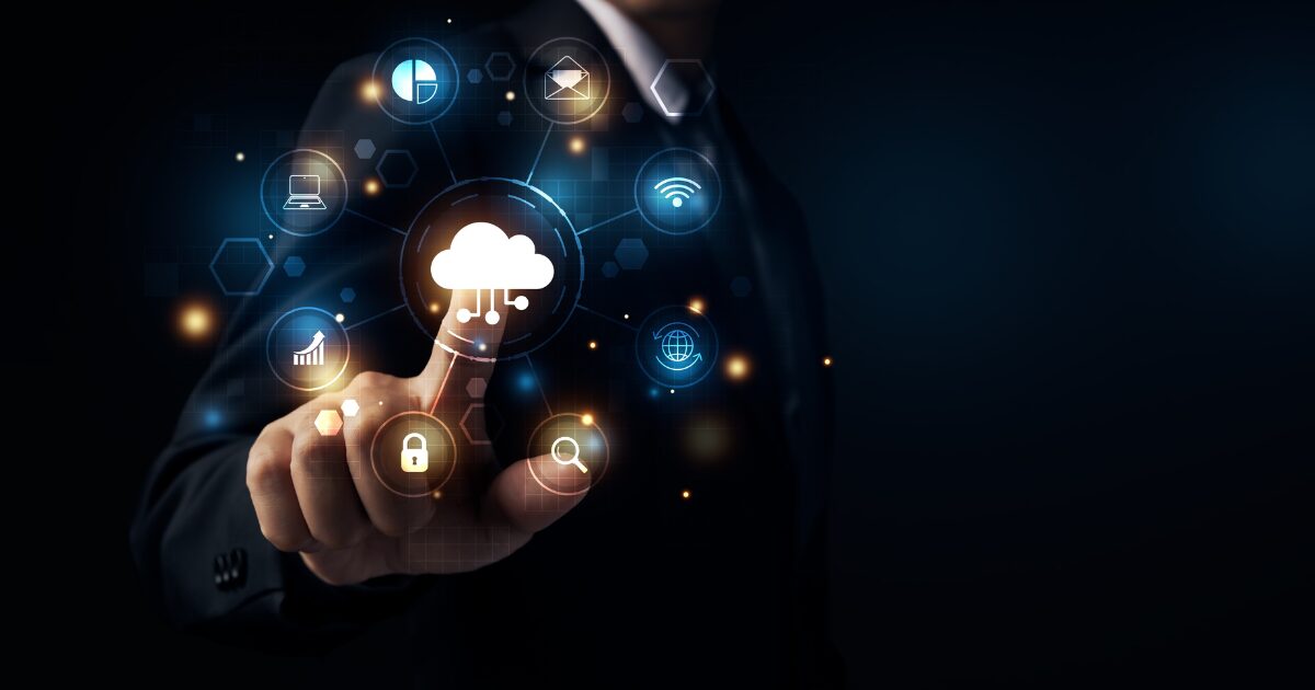 A imagem retrata uma mão de um profissional em traje formal, com o dedo indicador apontando para um ícone de nuvem iluminado em um fundo escuro. Ao redor do ícone da nuvem, há uma série de outros símbolos digitais que representam diferentes aspectos da tecnologia e da segurança da informação, como cadeados, engrenagens e gráficos. A cena é futurista e enfatiza a conexão com a computação em nuvem, sugerindo inovação e integração tecnológica no ambiente de negócios. A iluminação destaca os elementos digitais, criando um contraste atraente com o fundo escuro (nuvem, computação em nuvem, cloud)