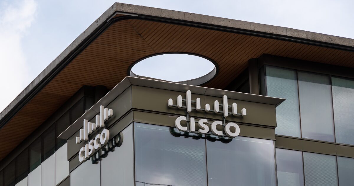 Imagem da fachada de um edifício da Cisco, empresa multinacional de tecnologia. O logotipo da Cisco está visível em duas laterais do prédio, com o nome da empresa em letras brancas e um ícone estilizado acima. A arquitetura moderna da construção conta com amplas janelas de vidro e um detalhe circular no teto. O céu ao fundo está nublado (colaboração, Nvidia)