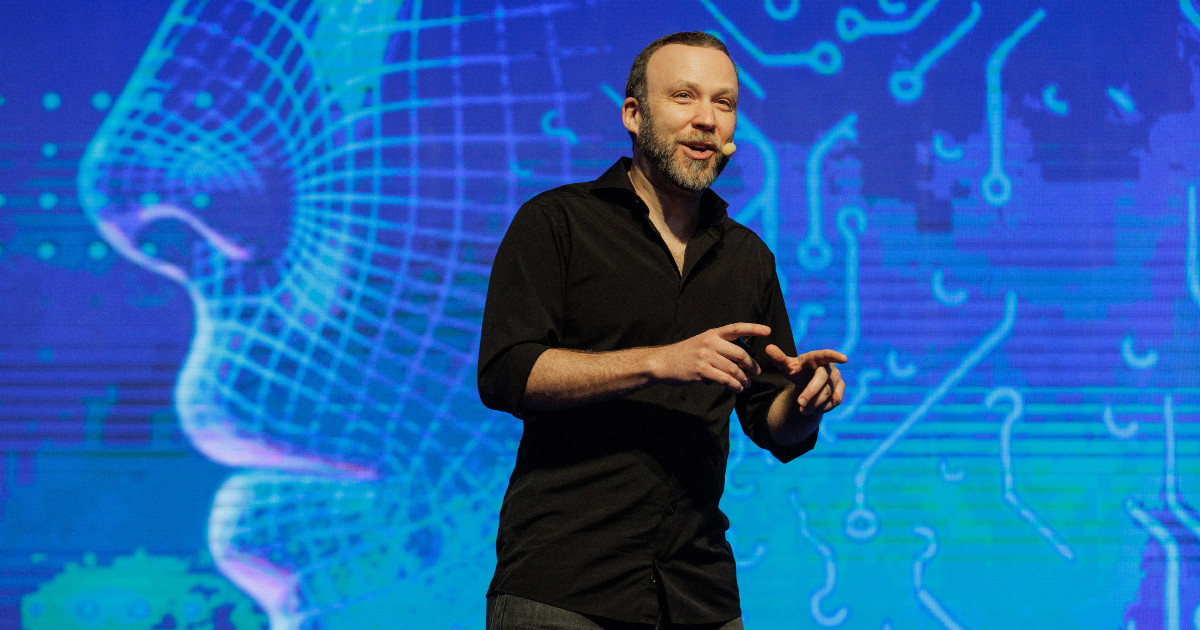 Dennis Herszkowicz, presidente da Totvs, durante uma apresentação no palco. Ele veste uma camisa preta e usa um microfone de headset enquanto gesticula ao falar. O fundo exibe uma grande projeção digital com elementos tecnológicos e a silhueta de um rosto, criando um ambiente futurista e inovador (Dennis Herszkowicz, Totvs, ERP)