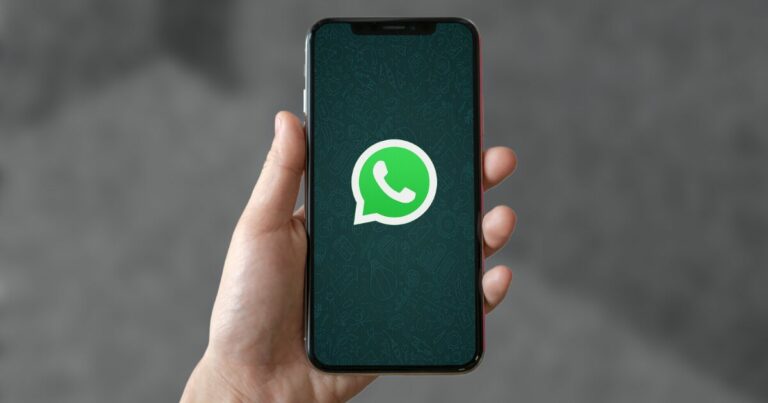 Imagem de uma mão segurando um smartphone que exibe a tela inicial do WhatsApp, com o ícone verde característico do aplicativo. O fundo desfocado em tons de cinza destaca o dispositivo e o logotipo do WhatsApp (whatsapp, mensagens, mensageria, varejo, rd station, Windows)