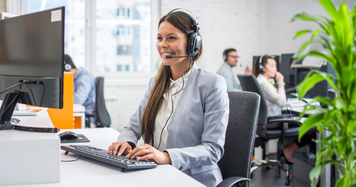 Mulher sorridente trabalhando em um escritório de call center, usando fone de ouvido com microfone, digitando no teclado de um computador. Ao fundo, outros colegas de trabalho estão atendendo chamadas em um ambiente corporativo iluminado e organizado, com plantas decorativas e janelas amplas que permitem a entrada de luz natural, nuvem