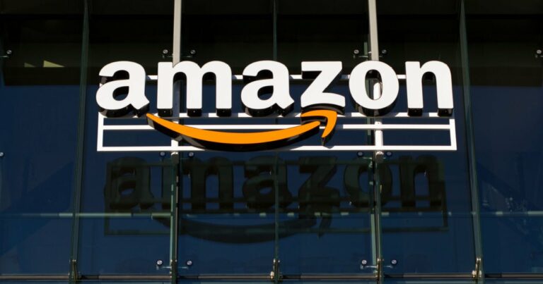 A imagem mostra o logotipo da Amazon em destaque, instalado na fachada de um prédio. O logotipo é composto pela palavra "amazon" em letras brancas, com a característica seta laranja que forma um sorriso abaixo da letra "a". A seta sugere a ideia de entrega rápida e satisfação. O fundo é formado por vidro reflexivo, o que permite que a imagem do logotipo seja refletida na superfície. A cena transmite um ambiente corporativo moderno e tecnológico (Amazon, dados, união europeia)