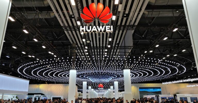 Estande da Huawei em um grande evento ou feira de tecnologia, com teto decorado por centenas de luzes em espiral e o logotipo da marca em destaque no centro superior da imagem. O local está lotado de pessoas, indicando grande interesse e movimentação no espaço da empresa. No fundo, é possível ver painéis e telas com informações tecnológicas, incluindo a frase "Accelerate industrial intelligence" (china, eua)