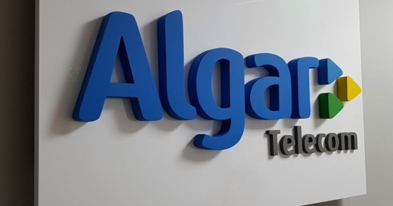 A imagem mostra a logo da Algar Telecom em um painel branco. O nome "Algar" está em letras tridimensionais azuis, enquanto "Telecom" aparece em letras menores e na cor preta. Ao lado do nome, há três setas coloridas apontando para a direita, nas cores azul, verde e amarela, que fazem parte da identidade visual da empresa, intern
