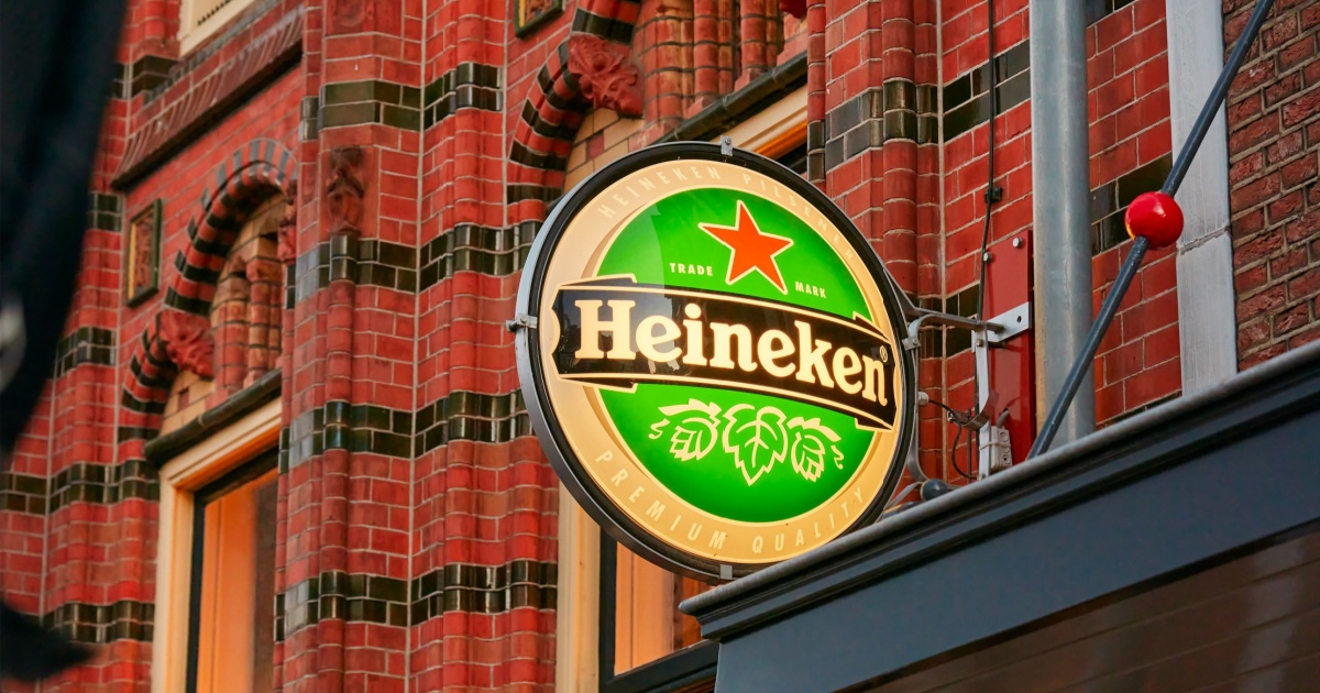 Placa luminosa redonda da marca de cerveja Heineken fixada na fachada de um prédio de tijolos vermelhos com detalhes em cerâmica verde e preta. A placa exibe o logotipo da Heineken em letras brancas sobre fundo verde, com uma estrela vermelha no topo e desenhos de lúpulo na parte inferior. Ao fundo, detalhes arquitetônicos decorativos do edifício.