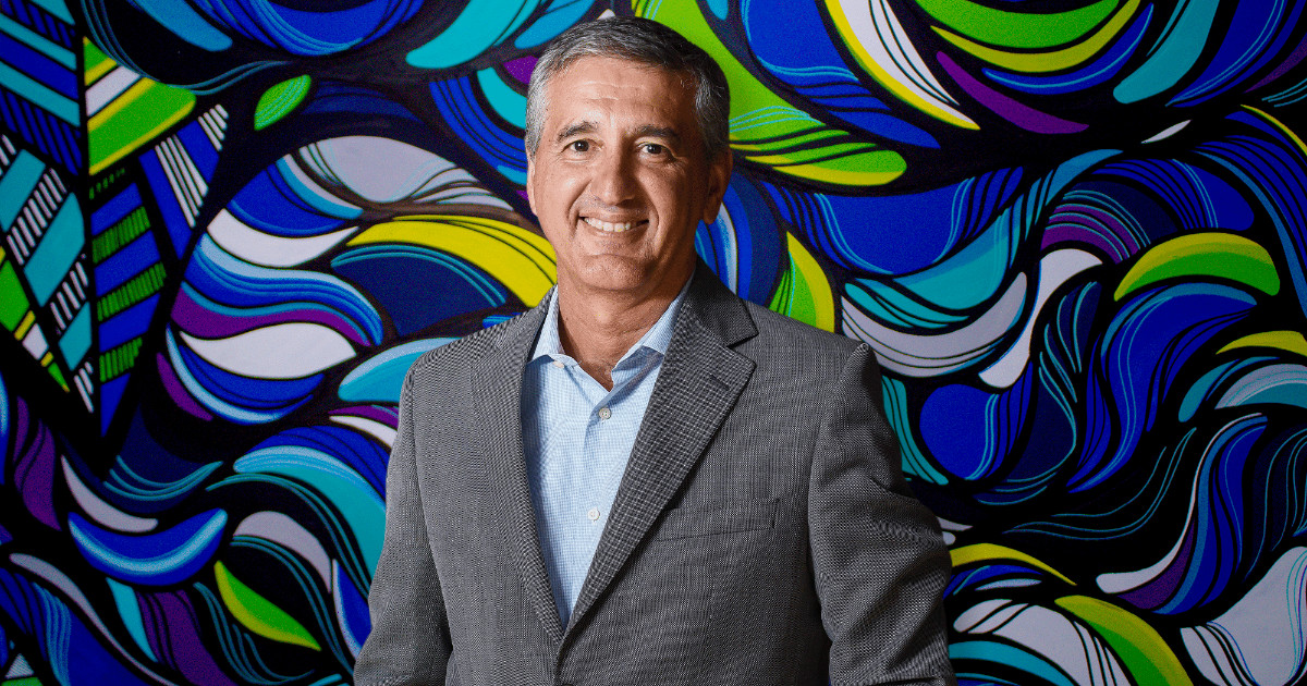 Marco Stefanini, fundador e CEO do Grupo Stefanini, sorrindo para a câmera em frente a um fundo vibrante e colorido com formas abstratas, transmitindo uma imagem de energia e criatividade. Ele veste um paletó cinza e camisa azul clara, destacando sua presença profissional