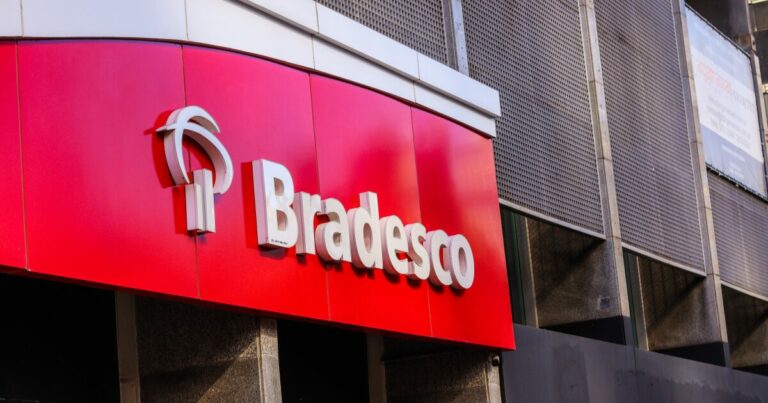 A imagem mostra a fachada de uma agência do banco Bradesco, com o logotipo característico em branco sobre um fundo vermelho. A estrutura tem um design moderno, com painéis metálicos perfurados ao fundo e um toldo curvo que abriga a identidade visual da instituição financeira (transações)