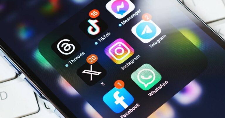 Tela de smartphone com vários aplicativos de redes sociais e mensagens abertos em uma pasta, incluindo Threads, TikTok, Messenger, Telegram, X (antigo Twitter), Instagram, Facebook e WhatsApp. Cada aplicativo exibe notificações de mensagens não lidas. A imagem está levemente inclinada, mostrando parte do teclado de um notebook ao fundo (apps, aplicativos, redes sociais, celular, smartphone, apps externos CGI, Windows)