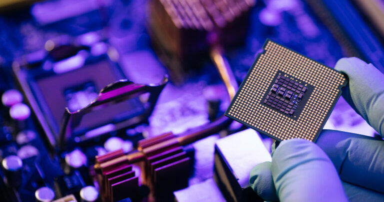 Imagem de close-up de um semicondutor sendo manuseado por uma pessoa usando luvas, com uma placa-mãe ao fundo. A cena destaca o componente essencial para o funcionamento de dispositivos eletrônicos (Chip, semicondutor, processador, chips, hardware, smicondutores, processadores, Intel, AMD, eletrônico, Gartner)