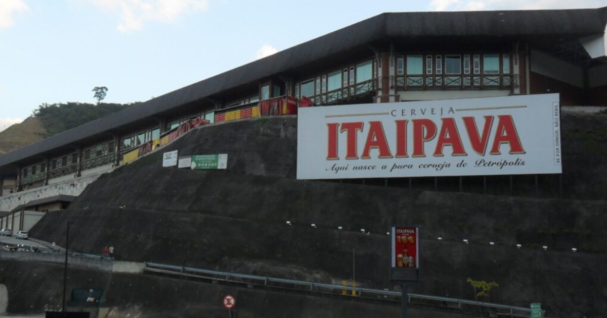 Imagem da fachada da fábrica da cervejaria Itaipava, localizada em Petrópolis, Rio de Janeiro. Em destaque, há um grande letreiro branco com letras vermelhas que diz: "Cerveja Itaipava – Aqui nasce a pura cerveja de Petrópolis". A estrutura do prédio apresenta arquitetura com elementos em madeira e vidro, e está posicionada em um terreno elevado à margem de uma estrada. (itaipava, grupo petropolis, Petrópolis, alunos)