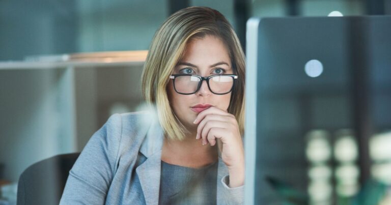 Mulher executiva de óculos, com expressão concentrada, analisando informações em uma tela de computador. Ela está em um ambiente de escritório moderno, vestindo um blazer cinza, transmitindo foco e profissionalismo (pensamento computacional, profissionais de TI, executiva, trabalho, mulher, trabalhadora, trabalhadores, profissionais, cresce)