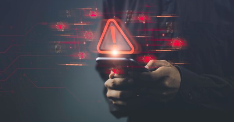 Pessoa segurando um smartphone com ícones e símbolos digitais em vermelho projetados no ar, incluindo um grande triângulo de alerta com um ponto de exclamação. A imagem simboliza um alerta de segurança cibernética ou vulnerabilidade em dispositivos móveis, com elementos gráficos digitais ao fundo (iot, ciberataque, vulnerabilidade, alerta, segurança, cibersegurança, dispositivo, device, golpe, ataques maliciosos)