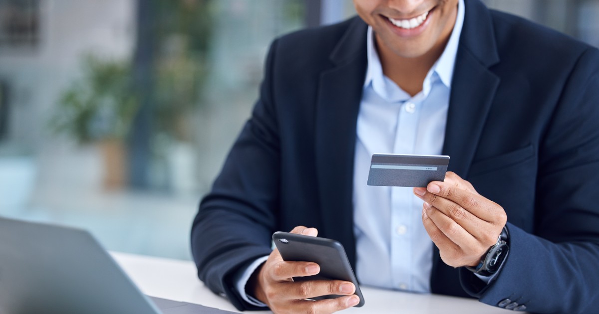 A imagem mostra um homem vestindo um terno escuro e camisa azul clara, segurando um cartão de crédito em uma mão e um smartphone na outra. Ele está sorrindo enquanto parece realizar uma transação ou compra online. No fundo, há um ambiente moderno e bem iluminado, possivelmente um escritório ou espaço corporativo. A cena transmite a ideia de tecnologia financeira, pagamentos digitais e transações seguras. (fintech, fintechs, crédito, dinheiro, cartão, brasileiros)