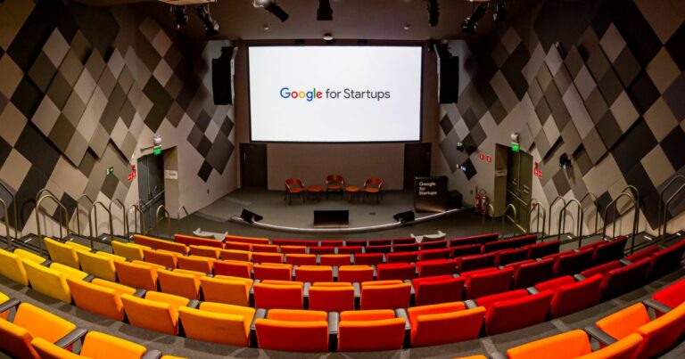 Auditório do Google for Startups, com assentos coloridos em tons de amarelo, laranja e vermelho, organizados em formato de arquibancada. Ao fundo, um grande telão exibe o logotipo 'Google for Startups', com cadeiras no palco para palestras. As paredes possuem design acústico geométrico (google for startups, google cloud)