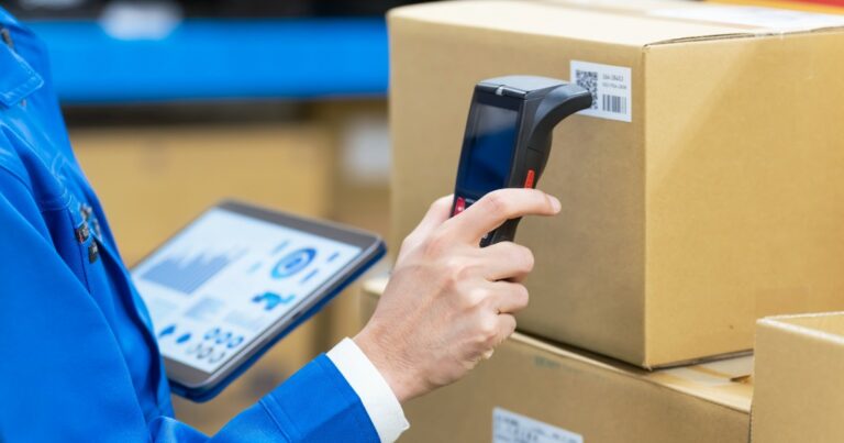 Trabalhador em um ambiente de logística utilizando um scanner de código de barras em uma caixa de papelão, enquanto segura um tablet com gráficos e dados exibidos na tela. Ele veste uniforme azul e está cercado por pacotes, sugerindo um processo de controle de estoque e rastreamento de produtos em um armazém (carbono)