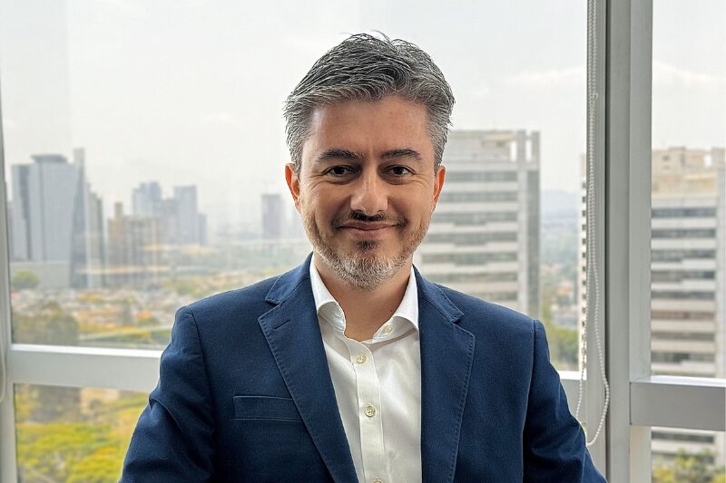 Retrato profissional de um homem de cabelos curtos e grisalhos, usando um terno azul e camisa branca. Ele está sorrindo levemente e posicionado em frente a uma janela com vista para prédios e uma paisagem urbana ao fundo, André Matos Arklok