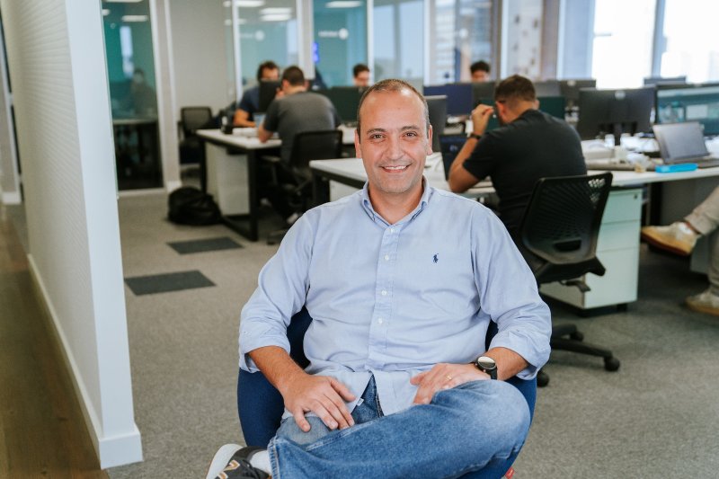 Foto de Giulliano Carvalho, CTO da Astride, sentado em uma cadeira em um ambiente de escritório moderno e descontraído. Ele veste uma camisa azul clara e jeans, sorrindo para a câmera. Ao fundo, outros colaboradores trabalham em mesas compartilhadas, criando uma atmosfera de produtividade e colaboração
