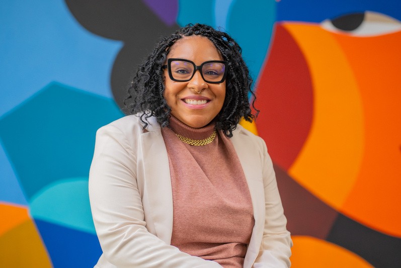 Cinthia Oliveira, head de ESG Global da CI&T, sorrindo em frente a um fundo vibrante e colorido com formas geométricas. Ela veste um blazer claro sobre uma blusa rosa e usa óculos de armação preta.