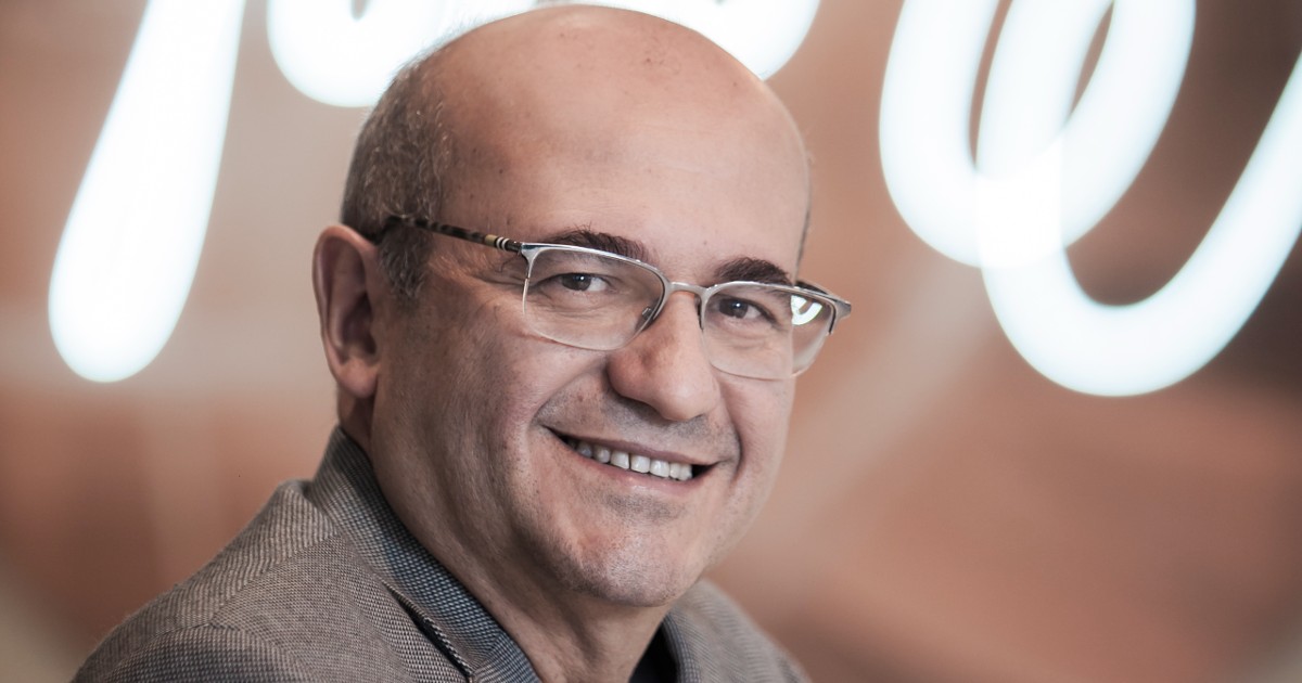 Cleber Morais, diretor-geral de vendas da AWS no Brasil, sorrindo para a câmera com um fundo desfocado em tons claros. Ele está usando óculos elegantes e um paletó cinza, transmitindo uma imagem de confiança e simpatia (IA generativa)