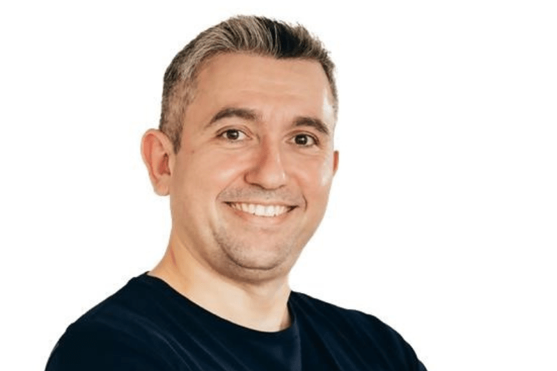 Retrato de um homem de cabelos curtos e grisalhos, sorrindo e olhando diretamente para a câmera. Ele veste uma camiseta escura e posa com os braços cruzados. O fundo é branco, destacando o rosto e a expressão acolhedora da pessoa, Daniel Moretto Nuvei