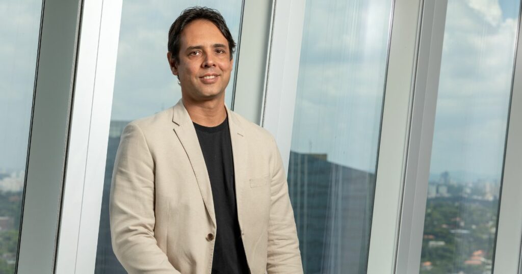 Emanuel Queiroz, executivo da Capgemini, posando em frente a uma ampla janela de vidro em um ambiente corporativo moderno. Ele veste um blazer bege sobre uma camiseta preta, com uma expressão confiante e o horizonte da cidade visível ao fundo