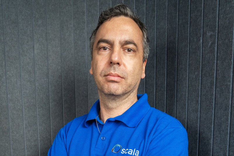 Foto de Filipe Cotait, CEO da Scala Stefanini, vestindo uma camisa polo azul com o logotipo da empresa. Ele tem uma expressão séria e está posicionado diante de um fundo cinza com textura, transmitindo uma imagem profissional e confiante