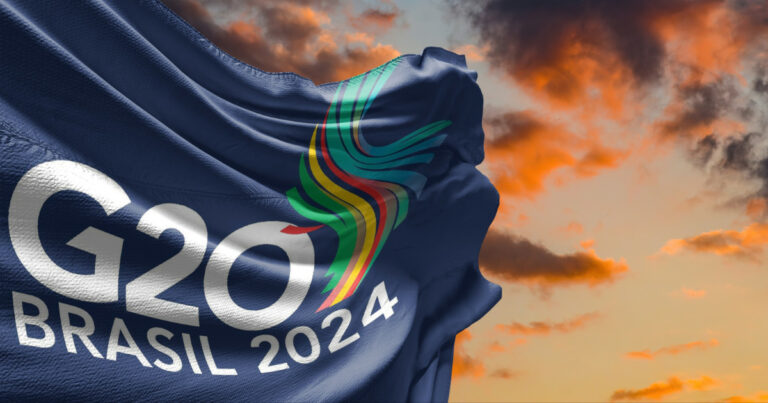 Imagem de uma bandeira azul escura com o logotipo do G20 Brasil 2024. O logotipo inclui a inscrição 'G20 Brasil 2024' em branco e um design colorido em forma de curva no centro, representando diversas cores vibrantes. Ao fundo, um céu com nuvens alaranjadas ao pôr do sol, criando um visual impactante e dinâmico (conservação ambiental)