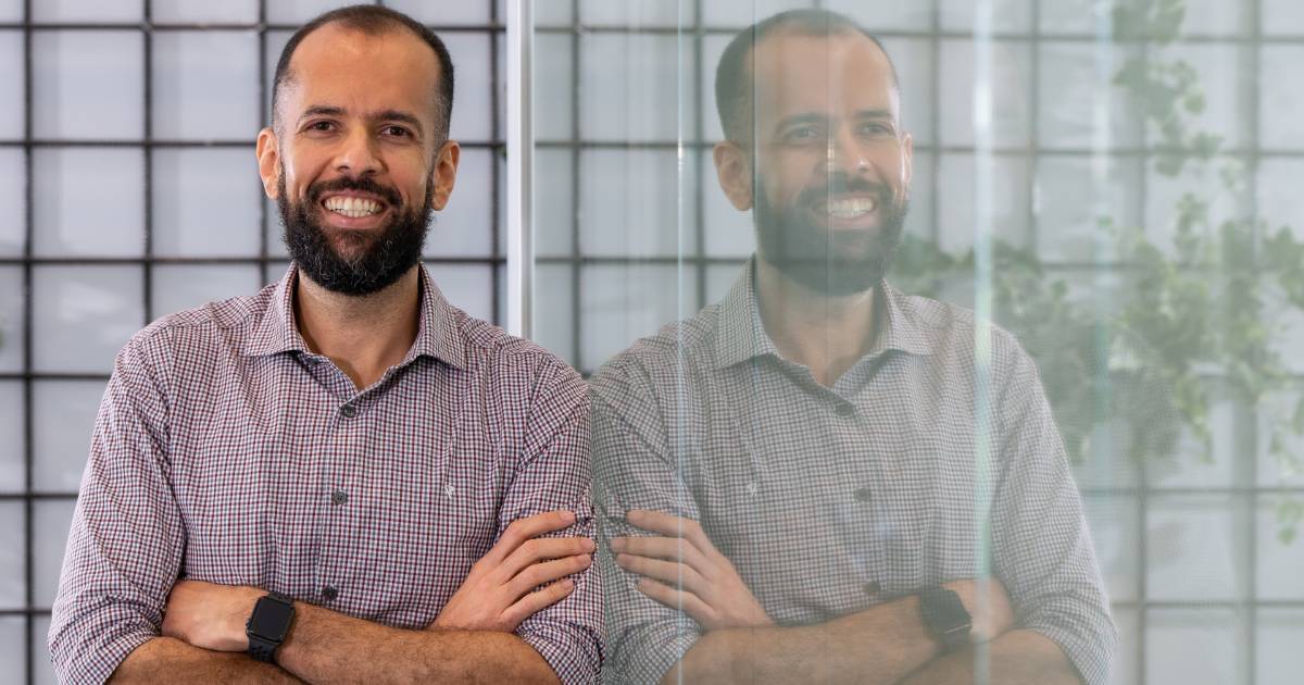 Gustavo Silva de França, diretor global de tecnologia e digital da Gerdau, sorrindo de braços cruzados ao lado de uma parede de vidro que reflete sua imagem. Ele veste uma camisa xadrez e usa um smartwatch, em um ambiente iluminado e moderno com detalhes de plantas ao fundo, conectividade