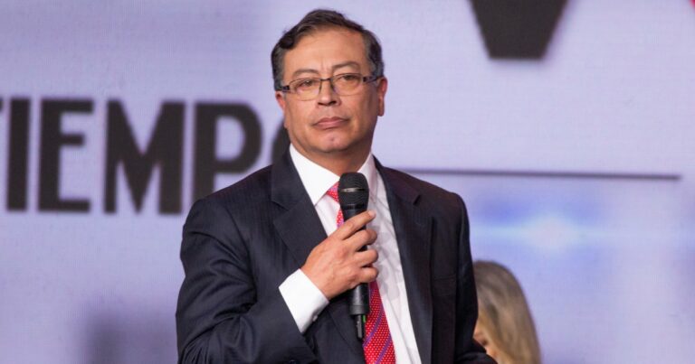 Foto de Gustavo Petro, presidente da Colômbia, em um evento público. Ele veste um terno escuro com gravata vermelha de bolinhas brancas e segura um microfone. Ao fundo, um painel desfocado com texto parcialmente visível, G20
