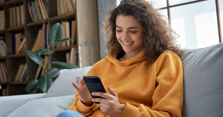 Jovem sorridente sentada em um sofá, usando um moletom amarelo e segurando um smartphone. Ao fundo, uma estante com livros e uma planta decorativa, em um ambiente bem iluminado e aconchegante. A imagem transmite conforto e conectividade, jovens