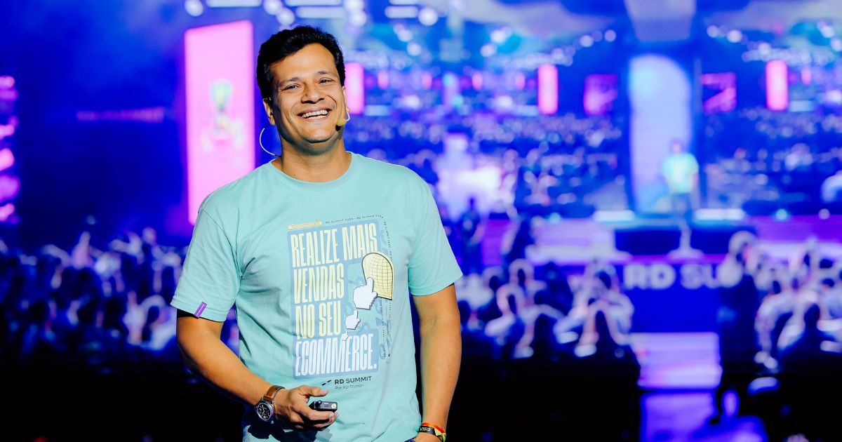 A imagem apresenta Juliano Tubino, CEO da RD Station, durante sua palestra no RD Summit. Ele está sorrindo e vestindo uma camiseta azul clara com uma mensagem que incentiva a realização de mais vendas no ecommerce. O fundo da imagem é vibrante, com um palco iluminado e uma plateia visível, destacando o ambiente animado e envolvente do evento. A atmosfera reflete a energia e a inovação que caracterizam a RD Station, especialmente em um contexto de crescimento e novas estratégias no mercado digital. ​​