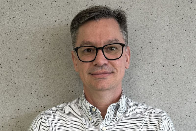 Retrato profissional de um homem de óculos, com cabelo curto e grisalho, vestindo uma camisa social clara com padrão discreto. Ele está posicionado em frente a uma parede de textura cinza, com uma expressão serena e confiante, olhando diretamente para a câmera, Marcio Facco, da Delfia
