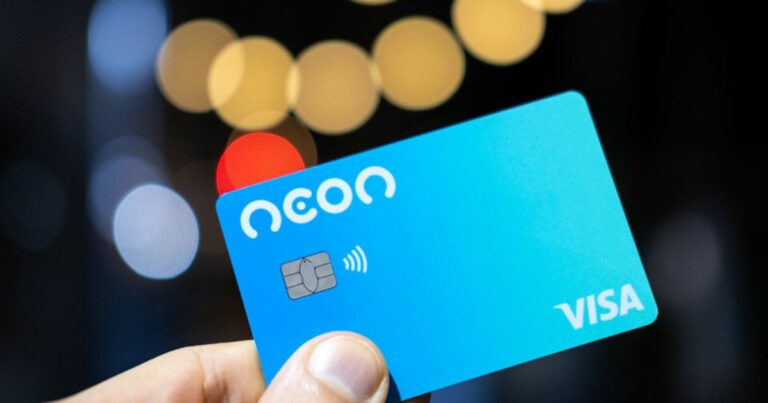 Imagem de um cartão Neon Visa azul, com design moderno e tecnologia de pagamento por aproximação, sendo segurado por uma mão. Ao fundo, um desfoque artístico com luzes em tons quentes, conferindo um visual sofisticado e contemporâneo